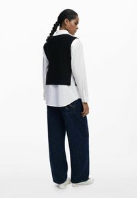Frau mit geflochtenem Haar, die ein weißes Langarmshirt unter einem schwarzen ärmellosen Pullover, weite dunkelblaue Jeans und weiße Schuhe trägt, steht seitlich.