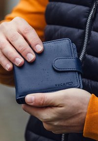 Navy leather wallet with a flap closure, textured surface, embossed logo, and visible water droplets. Held in a hand against a neutral background.