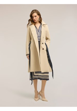 Donna che indossa un trench beige sopra un vestito a righe con motivi blu navy e bianchi, tacchi alti beige e grandi orecchini d'oro.