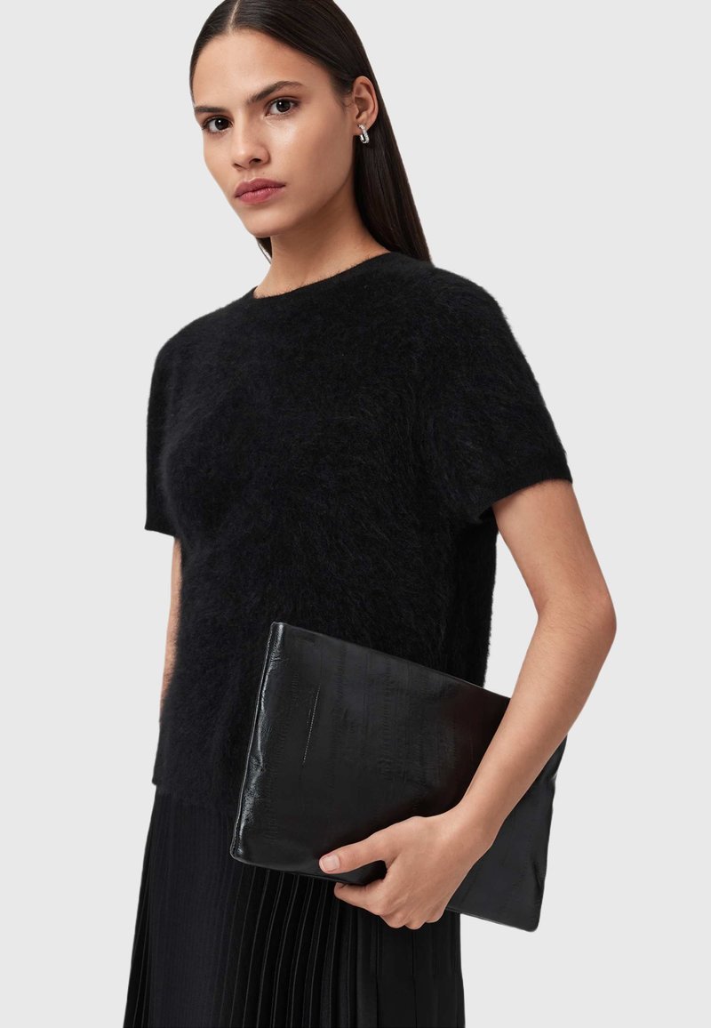 Maglione nero con texture e maniche corte, abbinato a una clutch nera con motivo a coccodrillo. Lo sfondo è neutro e minimalista.