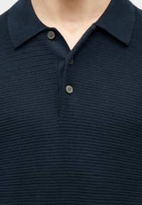 Κοντινή λήψη σκούρου μπλε πλεκτού πόλο πουκάμισου με τρία κουμπιά, ανοιχτό γιακά και ορατό περίγραμμα λαιμού.