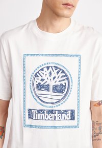 Valge puuvillane T-särk, millel on sinine graafiline disain puu logoga ja "Timberland" tekst ristkülikukujulises raamistikus. Lühikesed varrukad.