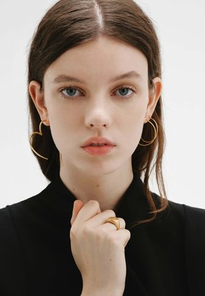 Joven con cabello oscuro que lleva pendientes de oro desiguales, uno en forma de corazón y otro grande en aro, además de un anillo de oro en la mano cerca del mentón.