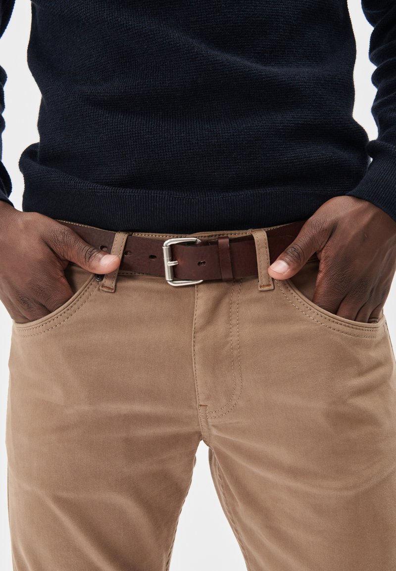 Rjave bombažne hlače z gladko teksturo, nošene z rjavim pasom z srebrno kovinsko sponko, v kombinaciji s temno modrim puloverjem.