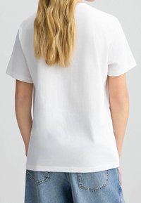 Valkoinen puuvillapaita, jossa on rento leikkaus, lyhyet hihat ja ympäri kaula-aukko. Sujuva pinta, helma päättyy juuri denim-shortsien ylle.