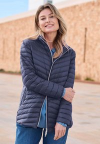 Marineblauwe pufferjack met diagonale gestikte ontwerpen, rits aan de voorkant, hoge kraag en elastische manchetten, gelaagd over een denimhemd.