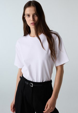 Donna con lunghi capelli castani che indossa una maglietta bianca semplice infilata in pantaloni neri a righe sottili con una cintura nera, in piedi davanti a uno sfondo chiaro.