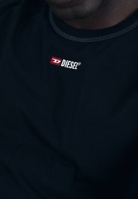 Black cotton t-shirt with a crew neck; features a red and white "DIESEL" logo on the upper left chest, contrasting stitching along the neckline.