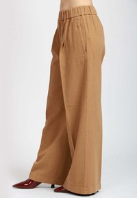 Pantaloni a gamba larga in un tessuto marrone chiaro con un finish testurizzato. Caratterizzati da una vita elastica e tasche laterali. Abbinati a tacchi a punta rossi.