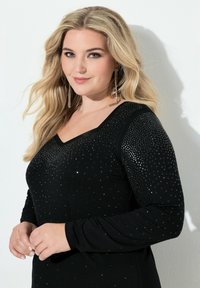 Svart långärmad topp med V-urringning, prydd med glittrande nitar över bål och axlar, med en figursydd siluett och texturerat tyg.