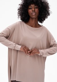 Licht taupe lange mouwen top met een ontspannen pasvorm, bootnek, zijsplitsingen en een gladde, zachte structuur. Geaccentueerd door een gebogen silhouet.