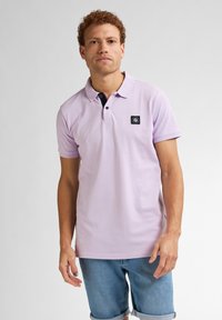 Světle fialová polo tričko s černým límečkem, dvoubotkovou légou a čtvercovým logem na levé hrudi. Krátké rukávy a texturovaná tkanina.