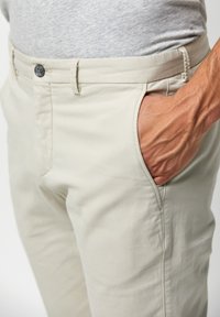 Chinos beige con una texture liscia, dotati di chiusura a bottone e tasche laterali. Una mano è appoggiata nella tasca, evidenziando un design aderente.