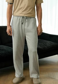 Man wearing beige T-shirt, light gray sweatpants with drawstring, and beige slippers standing on wooden floor near black sofa.