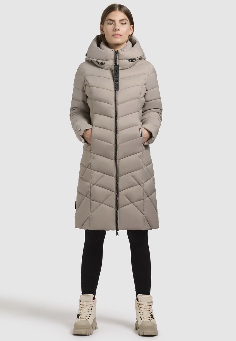 Podložena jakna v svetlo bež barvi z kapuco, diagonalo kvadratnim vzorcem, sprednjim zadrgo in stranskimi žepi. Nosi se čez črne legice in škornje.