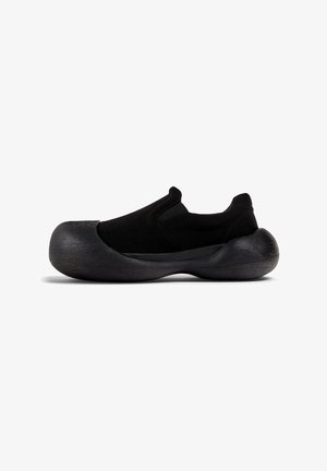 Zapatos negros sin cordones con parte superior de tejido y suela gruesa y texturizada. El diseño presenta una forma aerodinámica y acentos minimalistas.