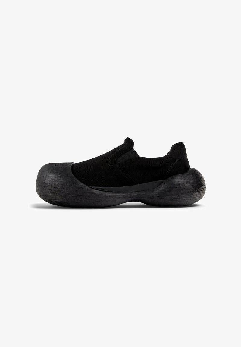Scarpe slip-on nere con tomaia in tessuto e suola spessa e texturizzata. Il design presenta una forma snella e accenti minimalisti.
