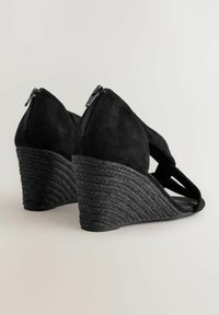 Sandales à talons compensés noires avec une tige en daim, présentant un design entrecroisé et un talon tissé texturé. Fermeture éclair à l'arrière.