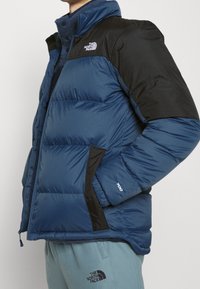 Blue puffy jacket featuring a black upper section, quilted texture, and logo accents. Includes zippered pockets and ribbed cuffs.