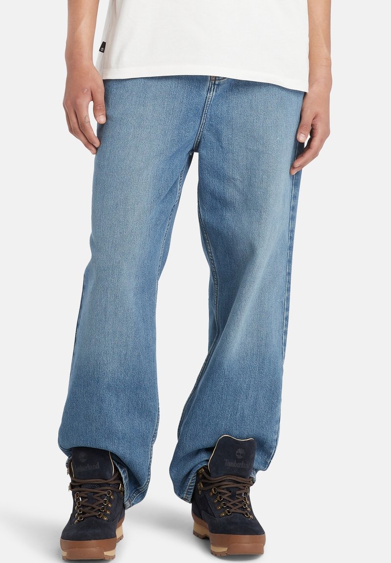 Personne portant un jean bleu clair ample, une chemise blanche, et des bottes Timberland marron foncé debout sur un fond blanc.