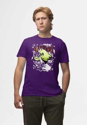 Lilla bomulds t-shirt med en tegneseriefigur i grøn med sorte detaljer, omgivet af hvide grafiske elementer og tekst. Korte ærmer.