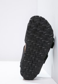 Musta sandaali, jossa on kuvioitu kumipohja ympyräkuviolla, korkkipohjallinen ja säädettävät remmit tukevan istuvuuden takaamiseksi.