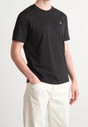 T-shirt en coton noir à manches courtes, avec un col rond et un petit logo sur la poitrine. Porté avec un pantalon de couleur ivoire.