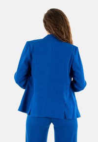Blazer azul con hombreras, corte ajustado y una abertura en la parte posterior. Hecho de tela suave, con un color sólido y líneas limpias.