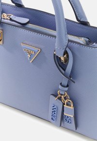 Lichtblauwe gestructureerde handtas met gouden accenten, voorzien van een driehoekig logo, ritssluiting en verwijderbare labels met merkinformatie.