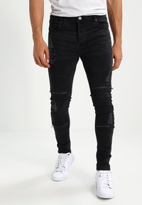 Hombre con jeans ajustados negros con cremalleras y parches desgastados, combinados con zapatillas blancas y una camisa blanca.