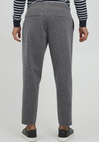 Graue Jogginghosen mit geradem Schnitt, ausgestattet mit zwei Gesäßtaschen, aus weichem Stoff. Kombiniert mit schwarzen Sneakers, die bis zu den Knöcheln sichtbar sind.
