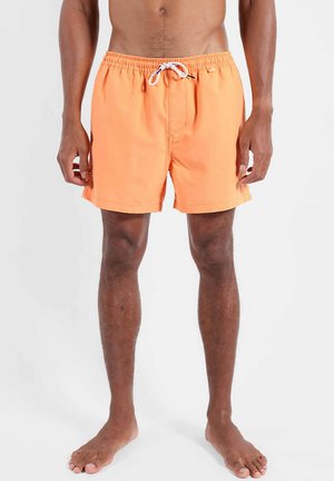 Buste et jambes masculins portant un short de bain orange vif avec un cordon blanc, debout pieds nus sur un fond clair uni.