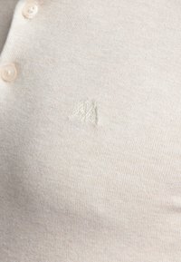 Ljust beige stickad polo skjorta med tre knappar, med en subtil broderad logotyp i samma färg. Mjuk, strukturerad tyg.
