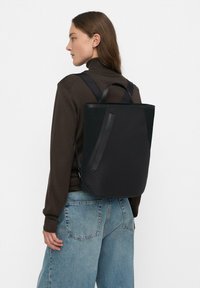 Czarny plecak o eleganckim designie, wykonany z gładkiego materiału, z siateczkowymi akcentami i dwoma górnymi uchwytami. Strukturalny, minimalistyczny kształt.