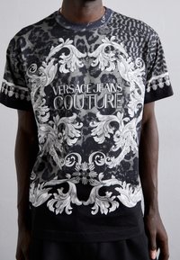 Svart t-shirt med vitt utsmyckat mönster och texten "VERSACE JEANS COUTURE", mot en texturerad djurtrycksbakgrund. Rund nacke, korta ärmar.