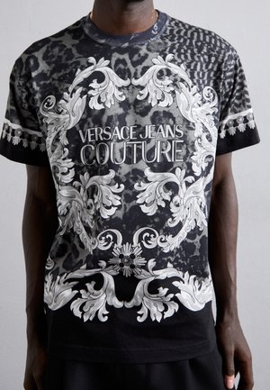 Schwarzes T-Shirt mit weißen, kunstvollen Mustern und dem Schriftzug "VERSACE JEANS COUTURE", vor einem strukturierten Tierdruck-Hintergrund. Runder Ausschnitt, kurze Ärmel.