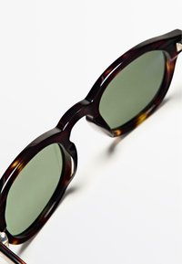 Zonnebril met schildpadmotief en ronde monturen, groene glazen en een glanzende afwerking. Voorzien van dikke pootjes en zilveren accenten bij de scharnieren.