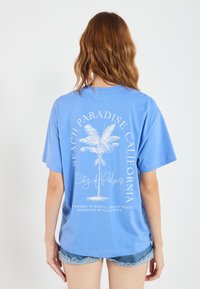 Light blue cotton t-shirt featuring a graphic of palm trees and text on the back. Short sleeves, relaxed fit, with raw edge detailing.