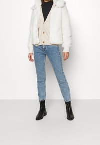 Bílá načechraná bunda s podšitým kapucí, přes béžový kardigan a černý top, doplněná světle modrými dž�íny a černými kotníkovými botami.