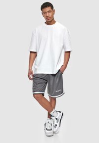 Bijela oversized majica u kombinaciji sa sivim sportskim šortsem s bijelim akcentima i logotipom. Model nosi crno-bijele tenisice sa čarapama.