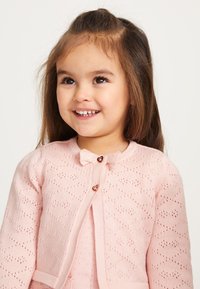 Ljusrosa stickad kavaj med mönstrad textur, hjärtformade knappar och en liten rosett vid kragen.