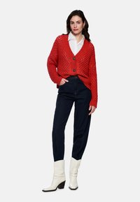 Roter, offen gestrickter Cardigan mit V-Ausschnitt, zwei vorderen Knöpfen, kombiniert mit dunklen Jeans und weißen, hochhackigen Stiefeletten. Dezente Texturdetails sind sichtbar.