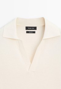 Katoenen poloshirt in crème met een textuurafwerking, voorzien van een klassieke kraag en een V-hals. Labels geven merk en materiaal aan.