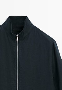 Veste noire à col montant avec fermeture éclair complète argentée à l'avant, présentée sur fond blanc.