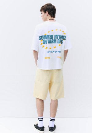 Ung mand stående med ryggen vendt iført en hvid t-shirt med blå og gul tekst og lysegule shorts mod en hvid baggrund.