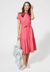 Koronkowa sukienka na guziki w kolorze koralowym, krótki rękaw i pasek w talii, z falbanistą spódnicą. Połączona z wzorzystą torbą na zakupy i białymi trampkami.