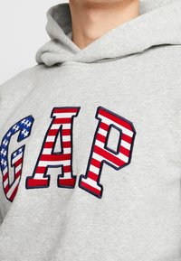 Sudadera gris con gran bordado de la palabra "GAP" en rayas rojas y blancas, con un pequeño patrón de estrellas azules en la letra 'G'.
