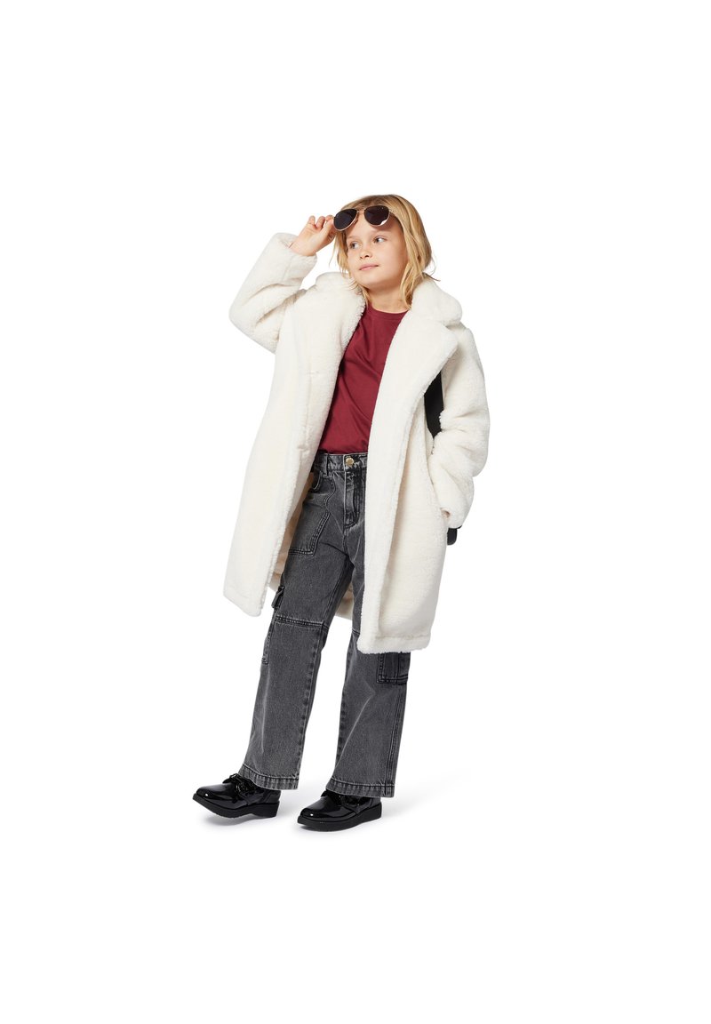Manteau en fausse fourrure blanche, chemise bordeaux, pantalon cargo gris et chaussures noires. Enfant portant des lunettes de soleil, prenant une pose.