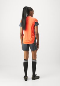 Camiseta atlética de color naranja brillante con detalles grises, combinada con pantalones cortos grises, calcetas negras hasta la rodilla con rayas blancas, y zapatillas de fútbol negras.
