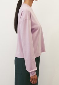 Light purple long-sleeve top with a relaxed fit, round neckline, and ribbed cuffs, paired with dark green trousers. Smooth texture.